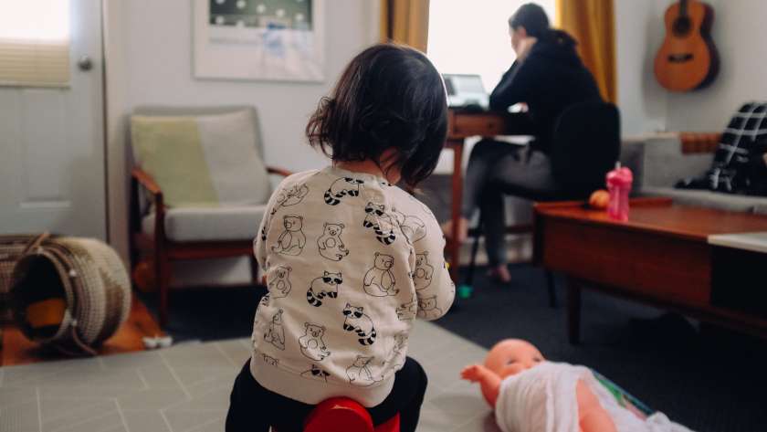 A toddler plays on the rug, surrounded by toys. In the background, a female figure (implied to be the mother) is sat at a desk, presumably working at a laptop.
