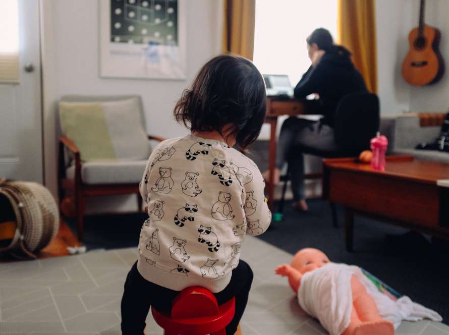 A toddler plays on the rug, surrounded by toys. In the background, a female figure (implied to be the mother) is sat at a desk, presumably working at a laptop.